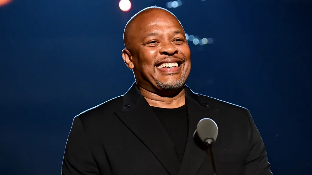 Dr. Dre on stage in a black suit jacket and vest, grinning from ear to ear, mic in hand